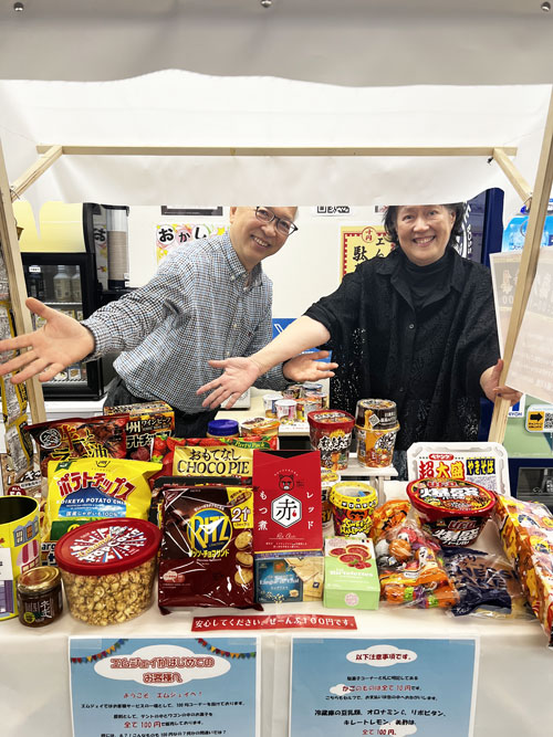 100均コーナーに並べられた商品を前にポーズをとる社長夫妻